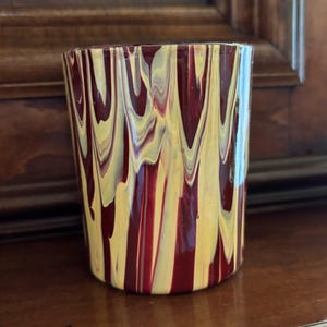 May include: A cylindrical glass with a distinctive design. The glass has a pattern of vertical streaks in shades of burgundy and yellow. The colours are swirled together, creating a visually interesting effect. The glass is on a wooden surface.