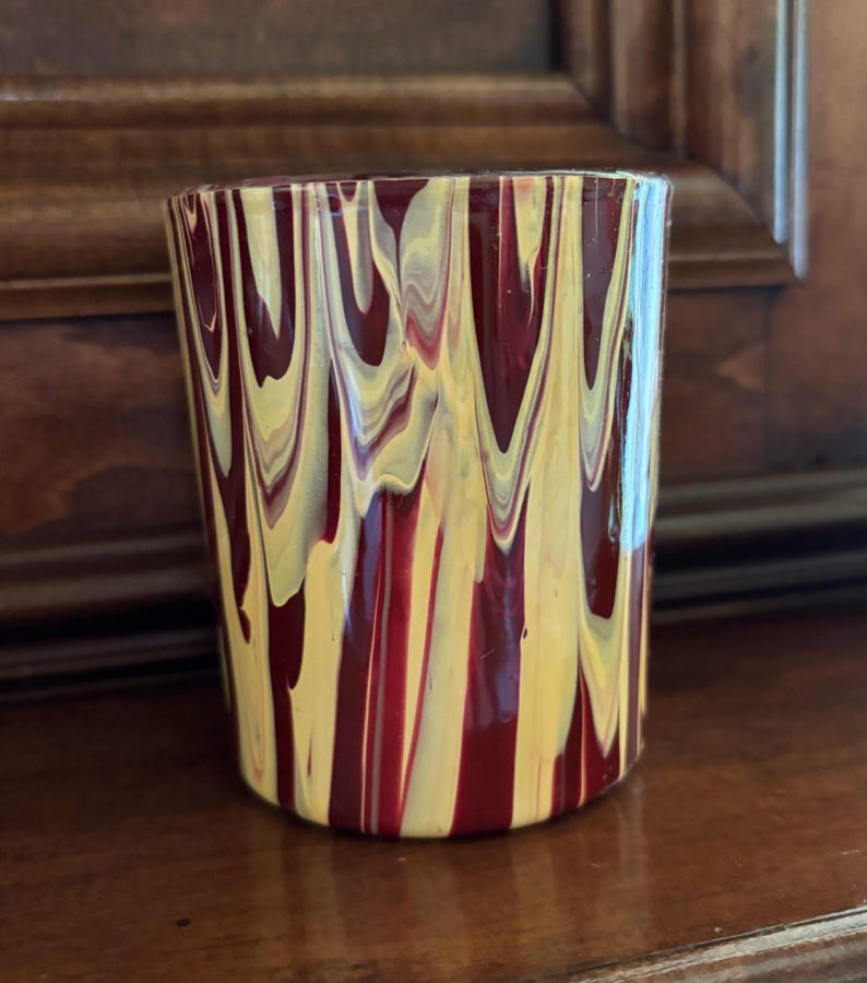 May include: A cylindrical glass with a distinctive design. The glass has a pattern of vertical streaks in shades of burgundy and yellow. The colours are swirled together, creating a visually interesting effect. The glass is on a wooden surface.