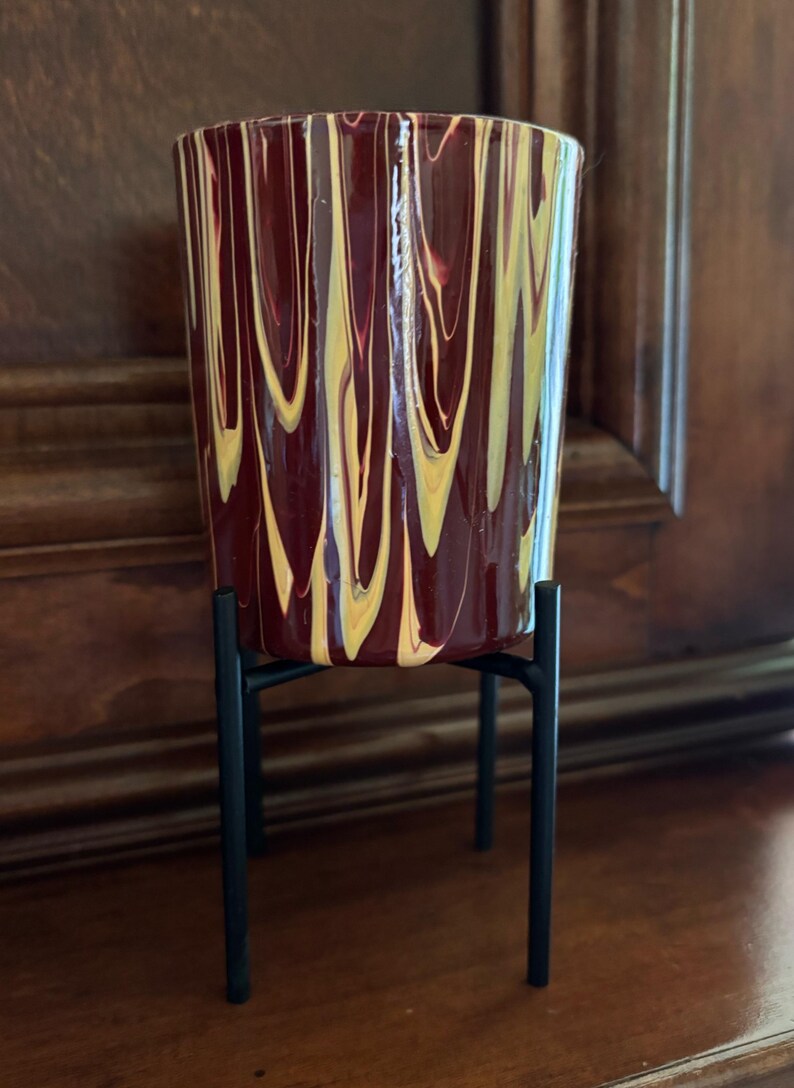 May include: A decorative cylindrical planter with a maroon and yellow swirled pattern, set on a black metal stand. The planter is approximately 15 cm tall and is suitable for indoor plants. The stand has a minimalist design.
