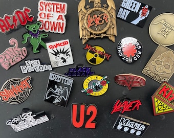 May include: A collection of enamel pins featuring various rock and metal bands. The pins include logos for bands like AC/DC, Slayer, Green Day, Metallica, and the Ramones. Other pins feature the Sex Pistols, Slipknot, and Gorillaz.