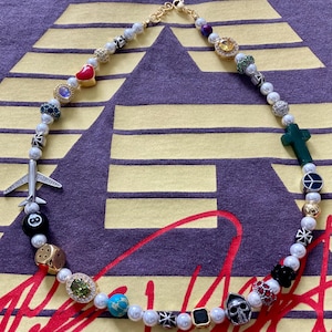 May include: A white pearl necklace with various charms, including a red heart, a green cross, a black and white dice, a black 8-ball, a turquoise stone, a gold skull, and a silver airplane.