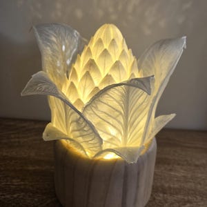 May include: A table lamp featuring a translucent white, artichoke-shaped shade with detailed leaf textures, illuminated from within by a warm light. The shade is mounted on a cylindrical, light-colored wooden base with a visible grain pattern.