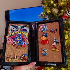 May include: A black display case with a corkboard interior, showcasing Disney pins. The left side features a large "Fantasia" pin with Mickey Mouse. The right side displays various character pins, including Mickey, Donald Duck, and a race car. A Christmas tree is in the background.