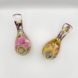 Op de afbeelding: Twee heldere glazen pijpen, elk gevuld met kleurrijke gedroogde bloemen. E&eacute;n pijp heeft roze en witte bloemen, terwijl de andere gele en paarse bloemen heeft. De pijpen hebben een druppelvorm met een gebogen mondstuk.