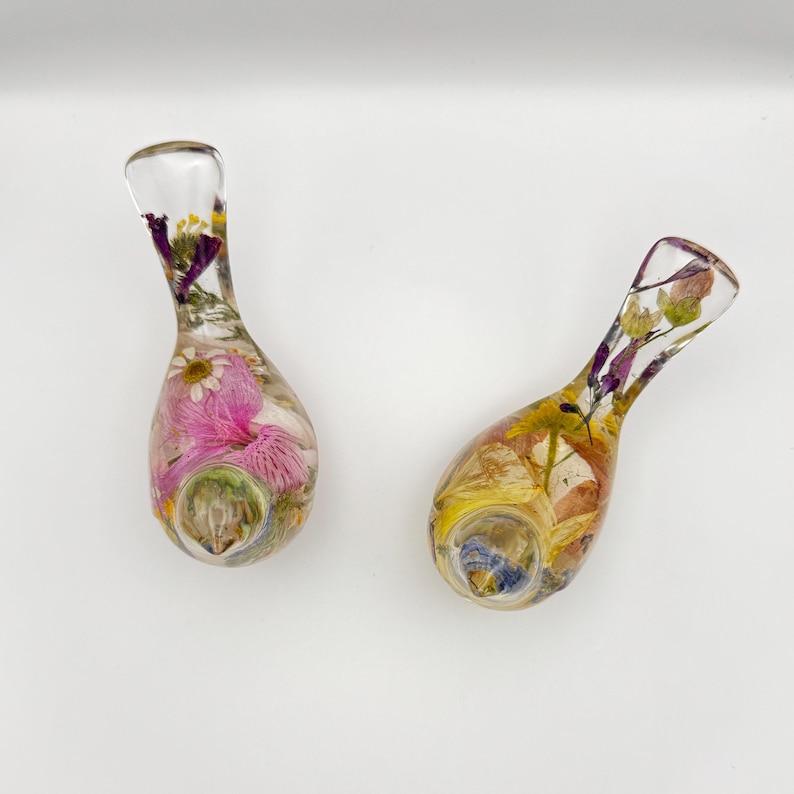 Op de afbeelding: Twee heldere glazen pijpen, elk gevuld met kleurrijke gedroogde bloemen. E&eacute;n pijp heeft roze en witte bloemen, terwijl de andere gele en paarse bloemen heeft. De pijpen hebben een druppelvorm met een gebogen mondstuk.