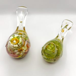 Op de afbeelding: Twee heldere glazen decoratieve stukken, elk met een bolvormige basis en een slanke hals. E&eacute;n stuk heeft een mix van gedroogde bloemen in tinten wit, geel en roze. De andere bevat groene bladeren en gouden accenten.