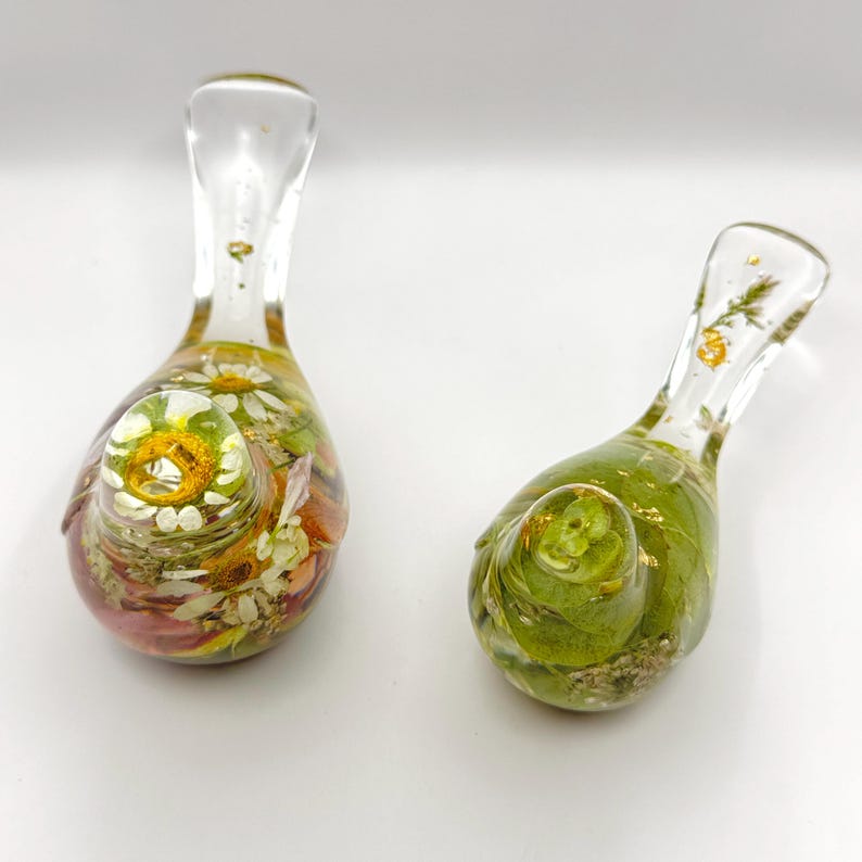 Op de afbeelding: Twee heldere glazen decoratieve stukken, elk met een bolvormige basis en een slanke hals. E&eacute;n stuk heeft een mix van gedroogde bloemen in tinten wit, geel en roze. De andere bevat groene bladeren en gouden accenten.