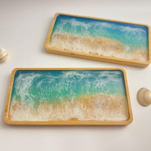 May include: Two rectangular wooden trays with a resin ocean design. The resin art features shades of blue and green, with white waves and a sandy beach effect. The trays are made of light-coloured wood.