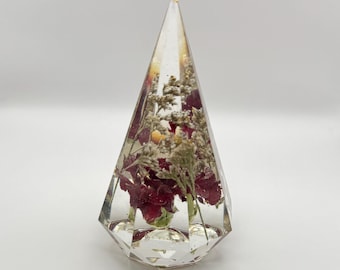 Op de afbeelding: Een heldere, facetgeslepen hars piramide met gedroogde rode bloemen en kleine witte en gele accenten. De piramide heeft een puntige top en een ruitvormige basis, waardoor een decoratief object ontstaat.