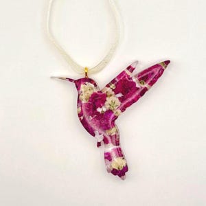 May include: Hummingbird pendant necklace with a white ribbon. The pendant is a hummingbird shape filled with pink and white flowers. The hummingbird pendant is approximately 3 inches long.