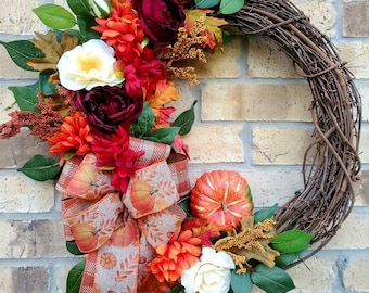 May include: A fall-themed wreath with a burlap bow, orange and white flowers, and a small orange pumpkin. The wreath is made of grapevine and is decorated with fall leaves.