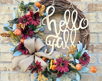 May include: A fall-themed wreath with a brown vine base, decorated with red and burgundy flowers, green leaves, and small orange pumpkins. The wreath features a wooden sign that reads "hello fall".