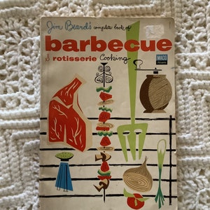 May include: Vintage cookbook titled "Jim Beard's complete book of Barbecue & Rotisserie Cooking." The cover features illustrations of barbecue tools, cuts of meat, and vegetables in red, green, blue, and brown. The book has a retro design with a mid-century modern aesthetic.