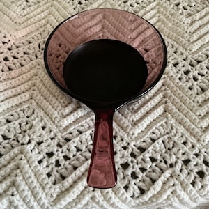 May include: A vintage, ruby-colored glass hand mirror with a round mirror and a long handle. The mirror has a dark, circular backing. The handle has a rectangular shape with a slightly flared base. The mirror is resting on a white, crocheted surface.