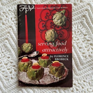 May include: Vintage cookbook cover titled "serving food attractively" by Florence Brobeck. The cover features artichokes, a red candle, and a serving dish with artichokes filled with red caviar and lemon slices. The background is a white crocheted blanket.