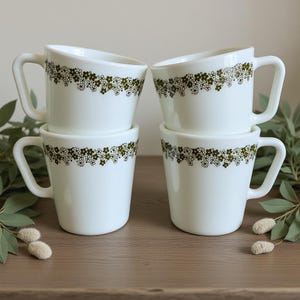 May include: Four white ceramic mugs with a green floral pattern around the top. Each mug has a curved handle and a slightly angled rim. The mugs are stacked in pairs on a wooden surface.