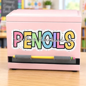 May include: A light pink rectangular pencil dispenser featuring a colorful sticker with the word PENCILS in large, multi-colored block letters and the script text Mrs. Likerland. A single yellow pencil rests horizontally in the dispenser slot.