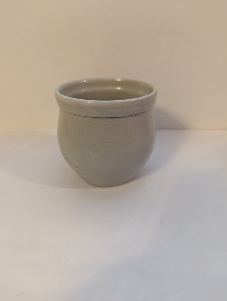 May include: A small, white ceramic cup with a rounded bottom and a slightly flared rim.