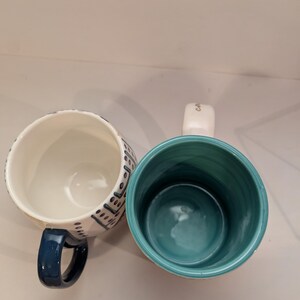May include: Two ceramic mugs with different designs. One mug has a white background with blue and black geometric patterns. The other mug has a white background with a teal blue interior and a gold inscription that reads "Coffee".