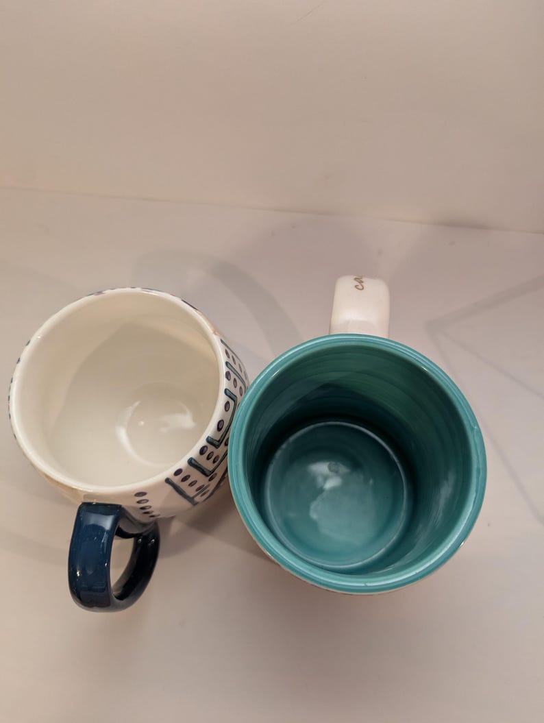 May include: Two ceramic mugs with different designs. One mug has a white background with blue and black geometric patterns. The other mug has a white background with a teal blue interior and a gold inscription that reads "Coffee".