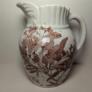 May include: A white ceramic pitcher with a brown floral design. The pitcher has a wide mouth and a handle.