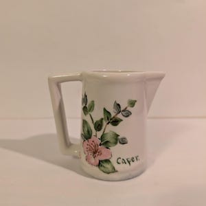 May include: A white ceramic creamer pitcher featuring a hand-painted floral design with a pink flower and green leaves. The word Caper is written in black script on the side. The pitcher has a square-shaped handle and a small pouring spout.