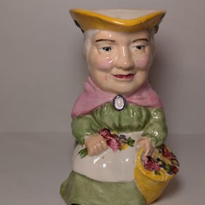 May include: A ceramic pitcher in the shape of a woman, wearing a yellow hat, pink shawl, and green dress. She holds a basket of flowers. The pitcher has a white apron and a smiling face.