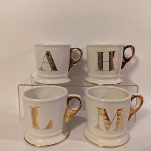 Pu&ograve; includere: Quattro tazze in ceramica bianca con bordo dorato e una lettera dorata sul davanti. Le lettere sono A, H, L e M.