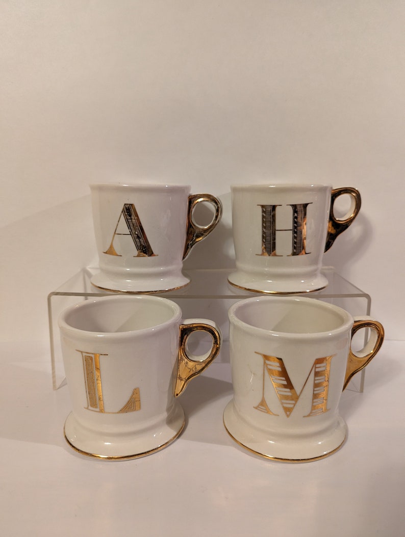 Pu&ograve; includere: Quattro tazze in ceramica bianca con bordo dorato e una lettera dorata sul davanti. Le lettere sono A, H, L e M.