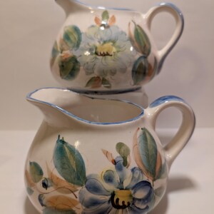 May include: Two ceramic cream pitchers with floral designs. The pitchers are white with blue rims and handles. Each pitcher features blue, green, and peach-colored floral patterns. One pitcher is stacked on top of the other.