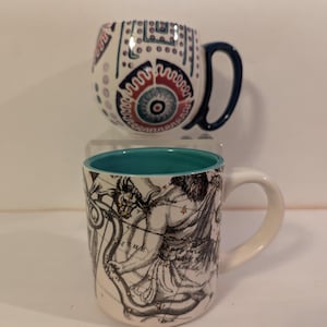 May include: Two ceramic mugs with different designs. The top mug has a white background with a blue, red, and white geometric pattern. The bottom mug has a white background with a black and white illustration of a constellation. The bottom mug has a teal blue rim.