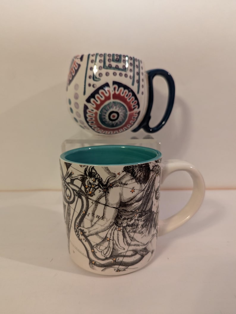 May include: Two ceramic mugs with different designs. The top mug has a white background with a blue, red, and white geometric pattern. The bottom mug has a white background with a black and white illustration of a constellation. The bottom mug has a teal blue rim.