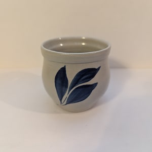 May include: A small, white ceramic mug with a blue leaf design. The mug is round and has a slightly flared lip.