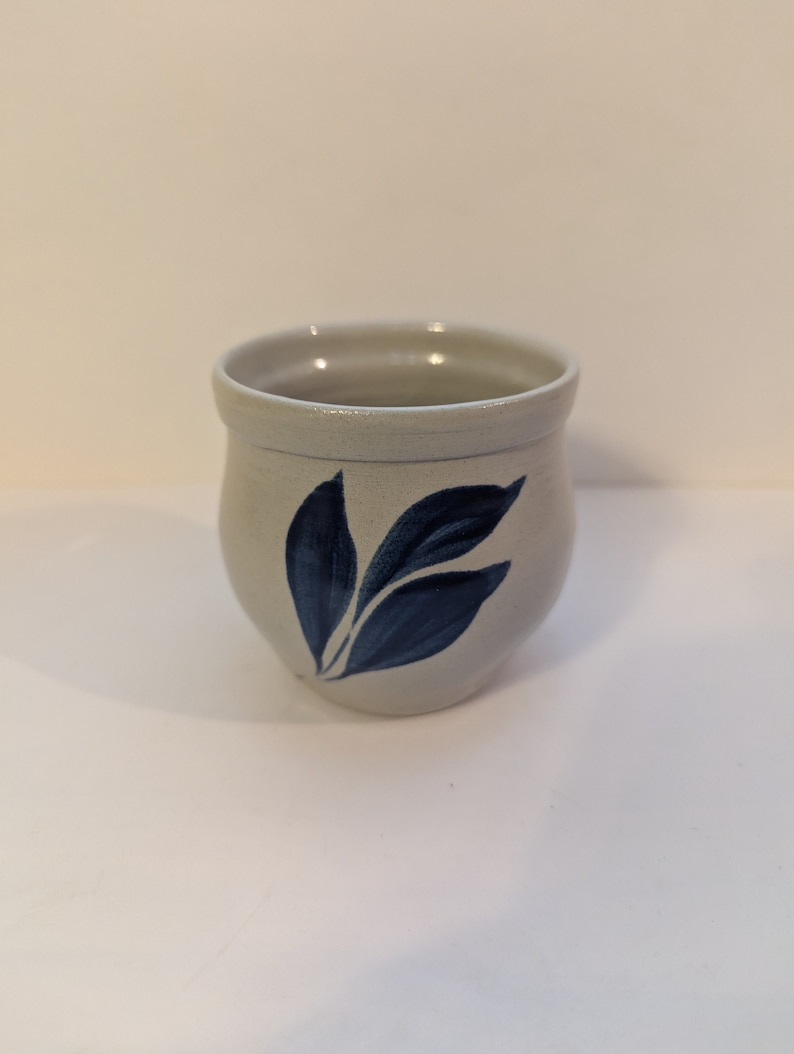May include: A small, white ceramic mug with a blue leaf design. The mug is round and has a slightly flared lip.