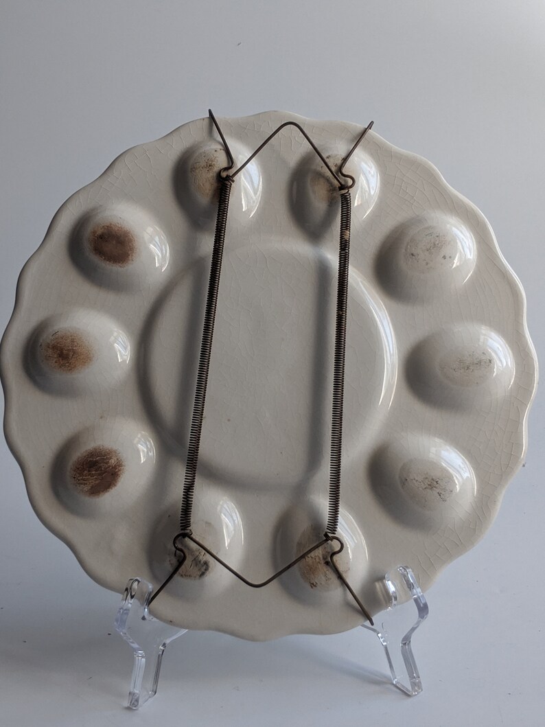 Puede incluir: Plato de huevos de cer&aacute;mica blanca con 10 hendiduras individuales para servir huevos. El plato tiene un borde festoneado y est&aacute; colgado de un soporte de alambre met&aacute;lico.