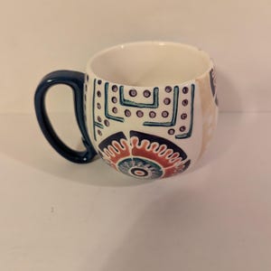May include: A white ceramic mug with a blue handle and colorful geometric patterns. The mug has a rounded shape and a wide base.