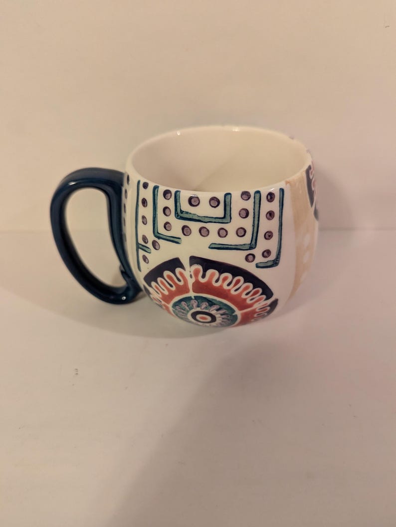May include: A white ceramic mug with a blue handle and colorful geometric patterns. The mug has a rounded shape and a wide base.