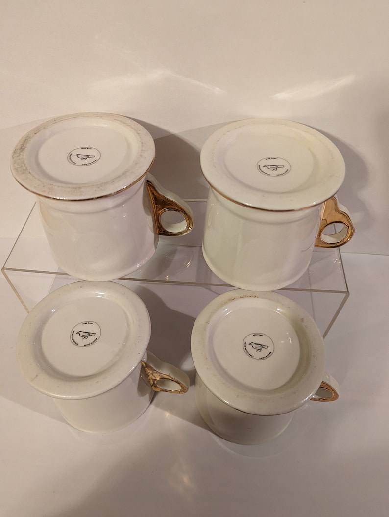 Pu&ograve; includere: Set di quattro tazze in ceramica bianca con bordo dorato e logo di un uccello dorato sul fondo.