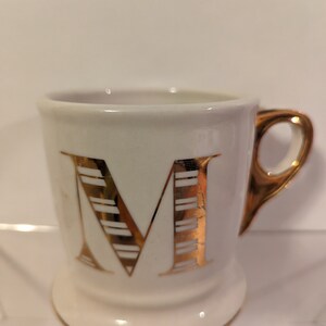 Pu&ograve; includere: Tazza in ceramica bianca con una lettera M dorata sul davanti. La lettera M ha un motivo a righe.