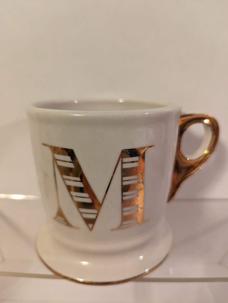 Pu&ograve; includere: Tazza in ceramica bianca con una lettera M dorata sul davanti. La lettera M ha un motivo a righe.