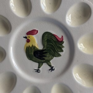Puede incluir: Un plato de huevos de cer&aacute;mica blanca con un dise&ntilde;o de gallo verde. El plato tiene ocho hendiduras individuales para sostener huevos.