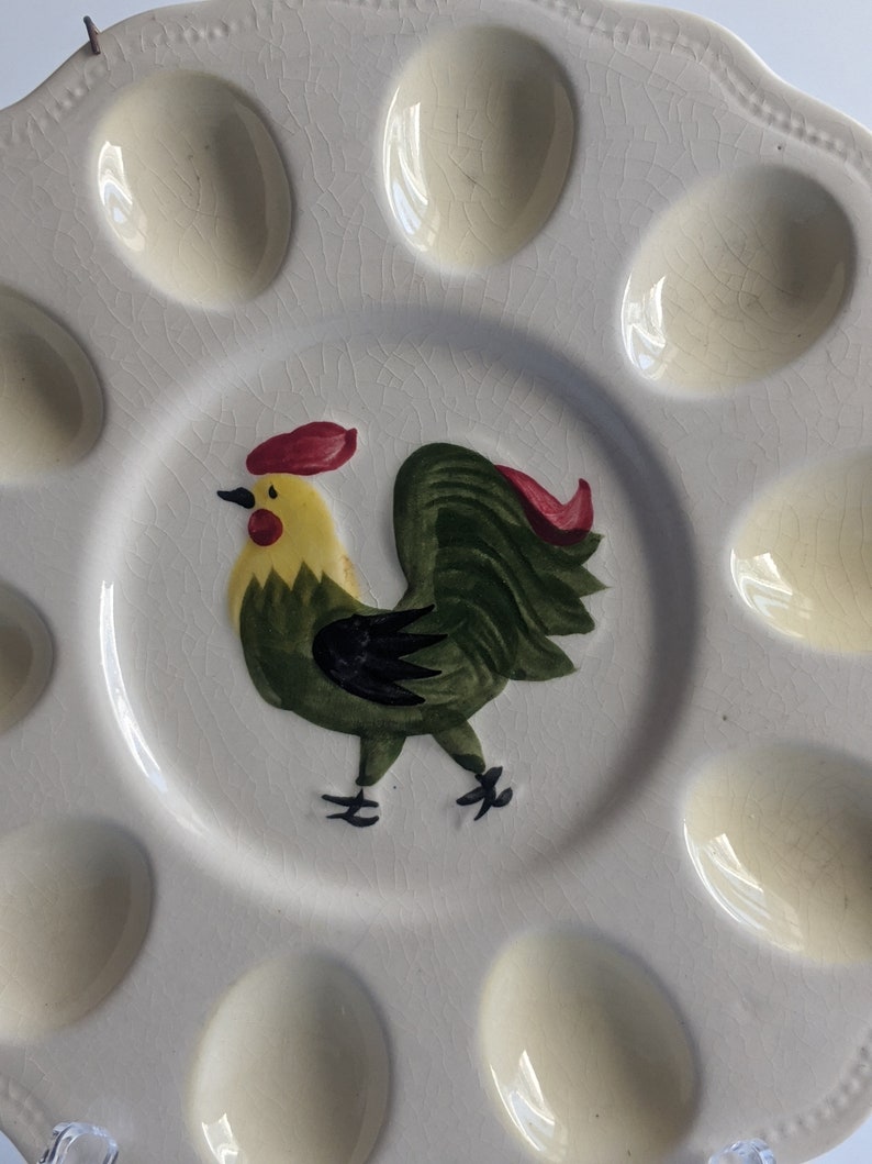 Puede incluir: Un plato de huevos de cer&aacute;mica blanca con un dise&ntilde;o de gallo verde. El plato tiene ocho hendiduras individuales para sostener huevos.