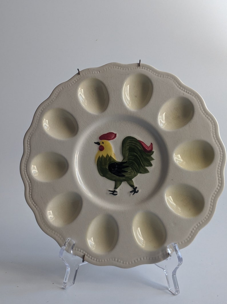 Puede incluir: Un plato de huevos de cer&aacute;mica blanca con 12 hendiduras individuales en forma de huevo. El plato presenta un gallo pintado a mano en verde, amarillo y rojo.