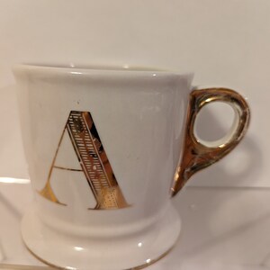 Pu&ograve; includere: Tazza in ceramica bianca con una lettera A dorata sul davanti e un manico dorato.