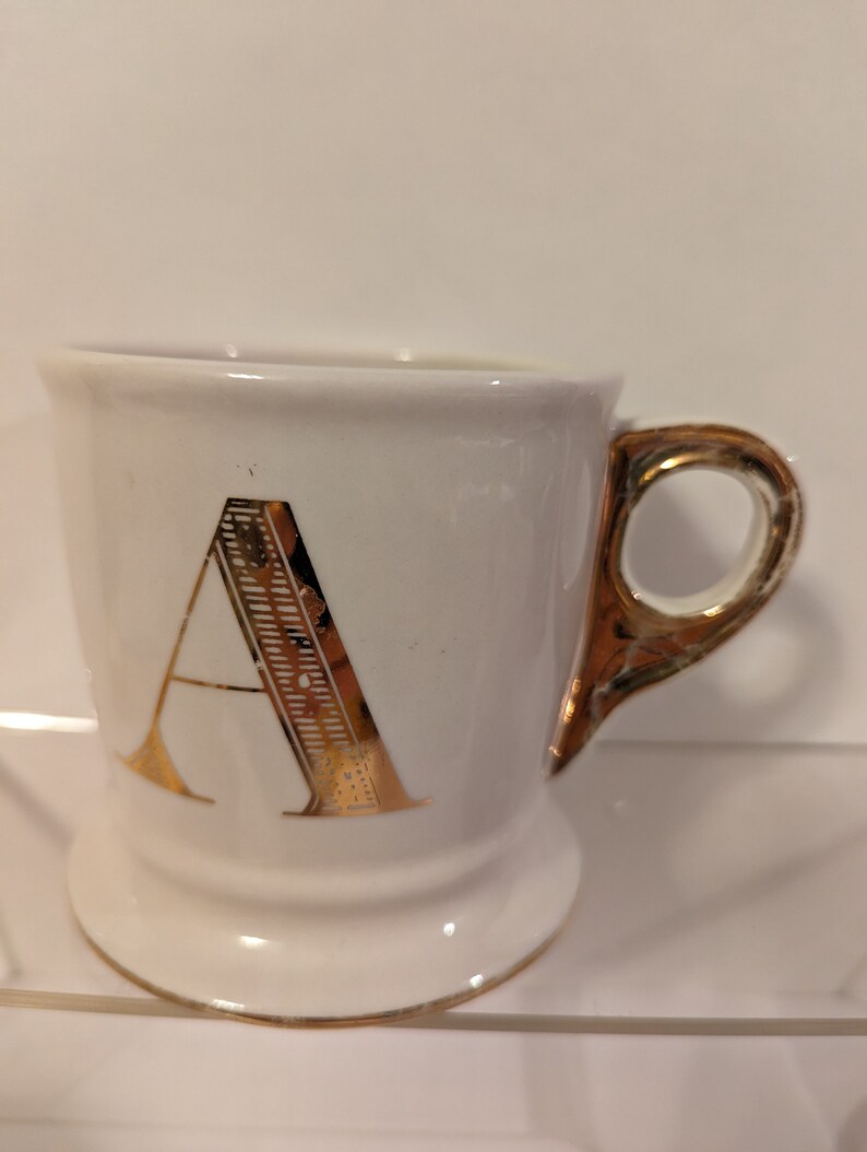 Pu&ograve; includere: Tazza in ceramica bianca con una lettera A dorata sul davanti e un manico dorato.