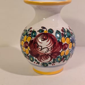 May include: A white ceramic vase with a rounded body and a flared rim. The surface features hand-painted floral motifs in shades of deep red, bright yellow, and teal green. The rim and base are accented with a solid yellow band and thin blue lines.