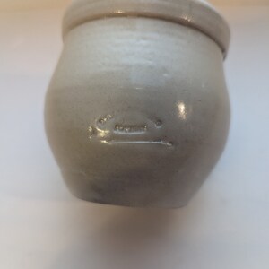 May include: A white ceramic crock with a rounded body and a slightly flared rim. The crock has a faint inscription on the front.