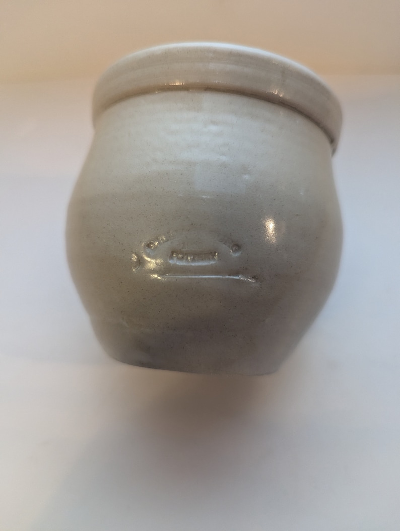 May include: A white ceramic crock with a rounded body and a slightly flared rim. The crock has a faint inscription on the front.