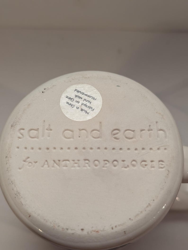 May include: White ceramic mug with the text "salt and earth for ANTHROPOLOGIE" on the bottom. The mug has a sticker on the bottom that reads "Made in China Fabriqu&eacute; en Chine hand wash recommended".