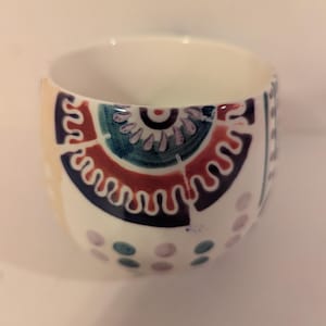 May include: A white ceramic cup with a colorful geometric design. The design features a large circle with a smaller circle inside, surrounded by a wavy line. The cup is decorated with dots and swirls in shades of blue, green, purple, and red.